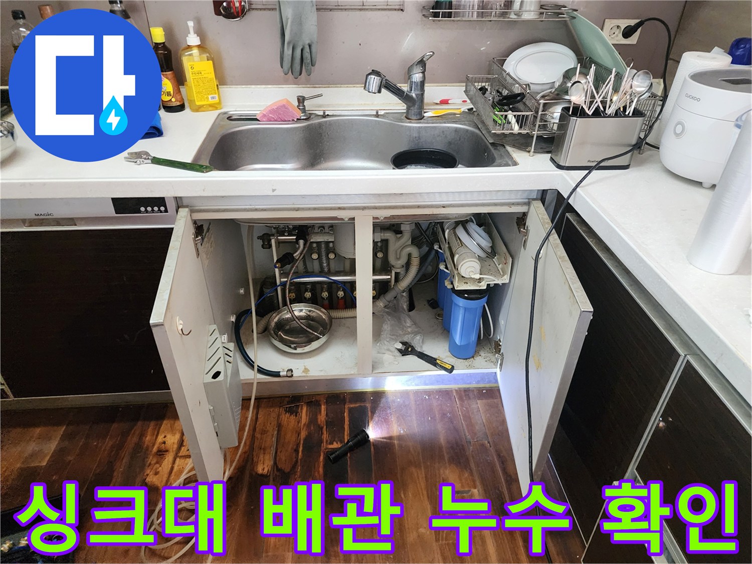 어두운 싱크대 하부장 내부의 배관 연결 부위를 손전등으로 밝게 비추는 모습