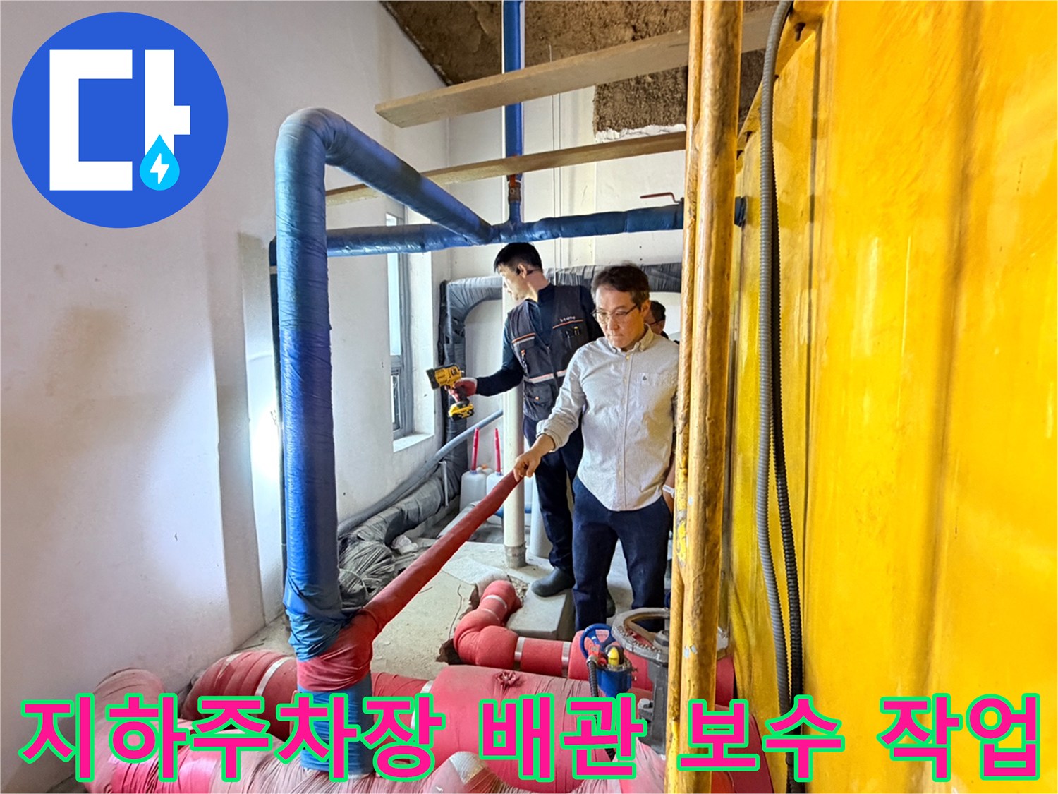 지하 주차장 천장의 파란색 및 분홍색 보온재로 감싸진 배관을 엔지니어가 점검하고 있습니다.
