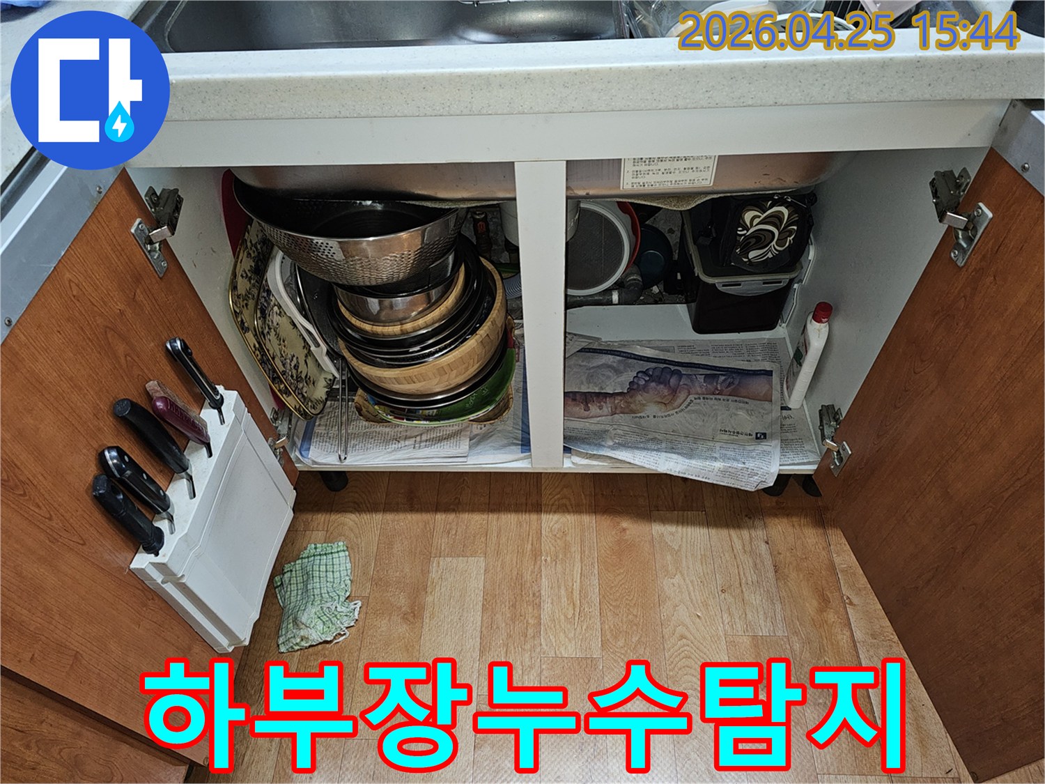 싱크대 하부장 내부-누수 점검을 위해-그릇과 배관이 보이고 바닥에 신문지가 깔려 있습니다.