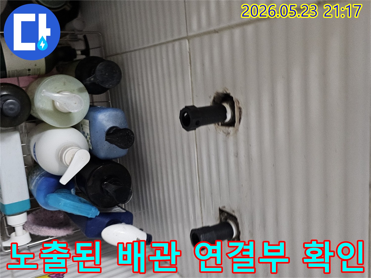 타일 벽면에서 튀어나온 두 개의 수도 배관 연결 부위를-누수 지점을 정확히 파악하기 위해-집중적으로 살펴보는 모습입니다.