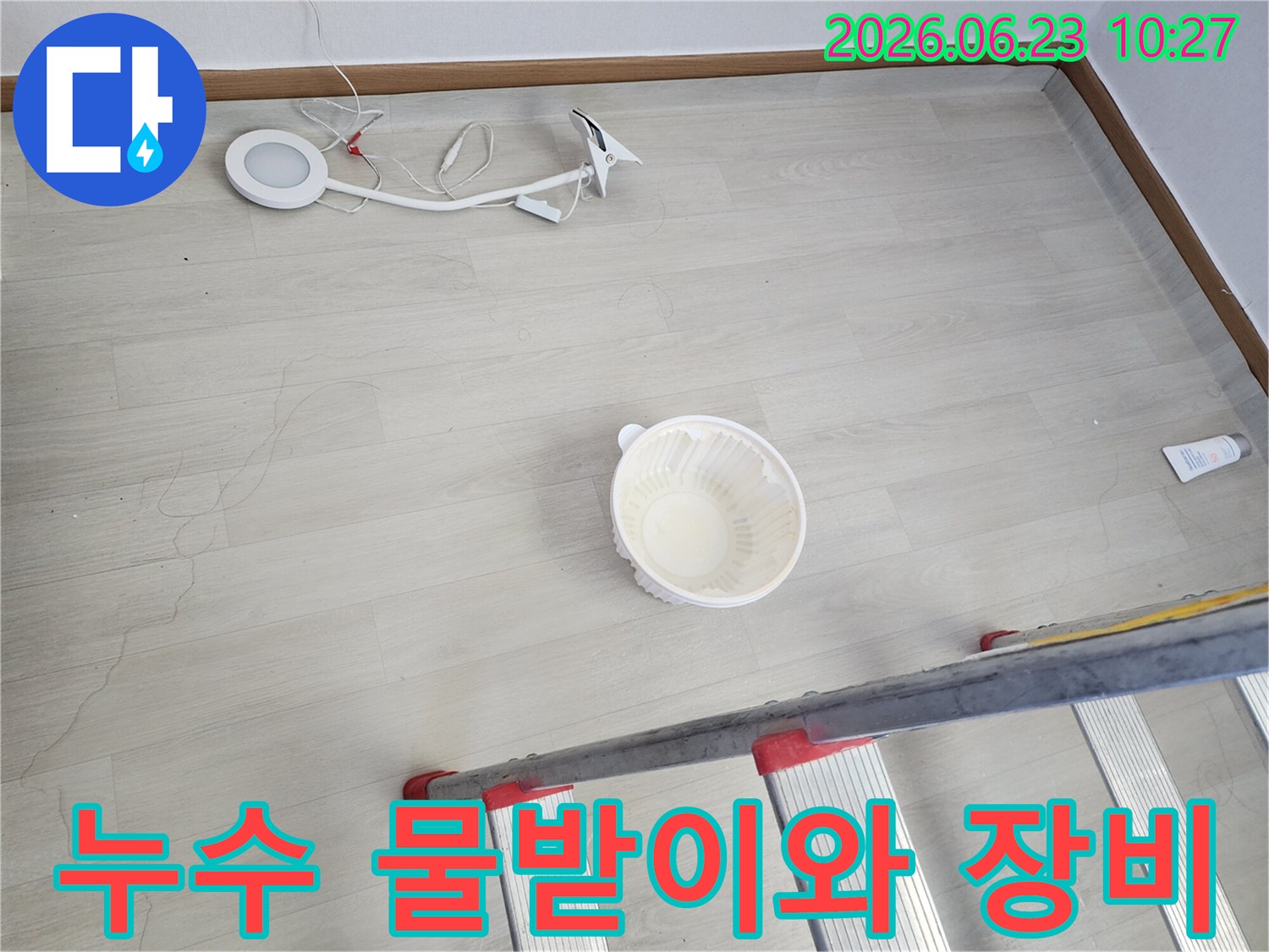 천장에서 물이 떨어져 바닥에 놓인 물받이 그릇과-누수 지점 확인 및 작업을 위한 사다리-조명 장비가 함께 보이는-누수 현장 준비 단계의 모습입니다