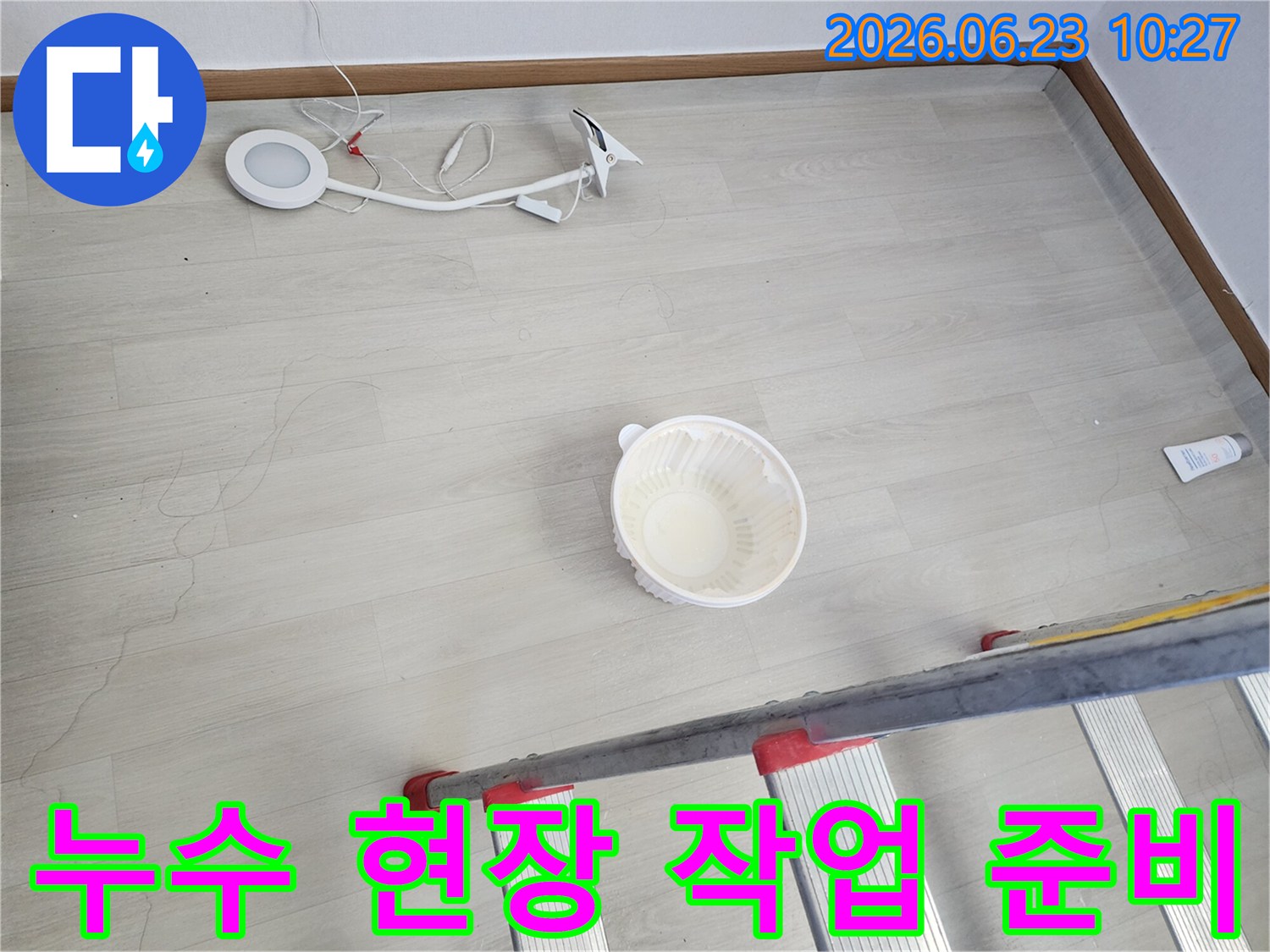 천장에서 물이 떨어져 바닥에 놓인 물받이 그릇과-누수 지점 확인 및 작업을 위한 사다리-조명 장비가 함께 보이는-누수 현장 준비 단계의 모습입니다