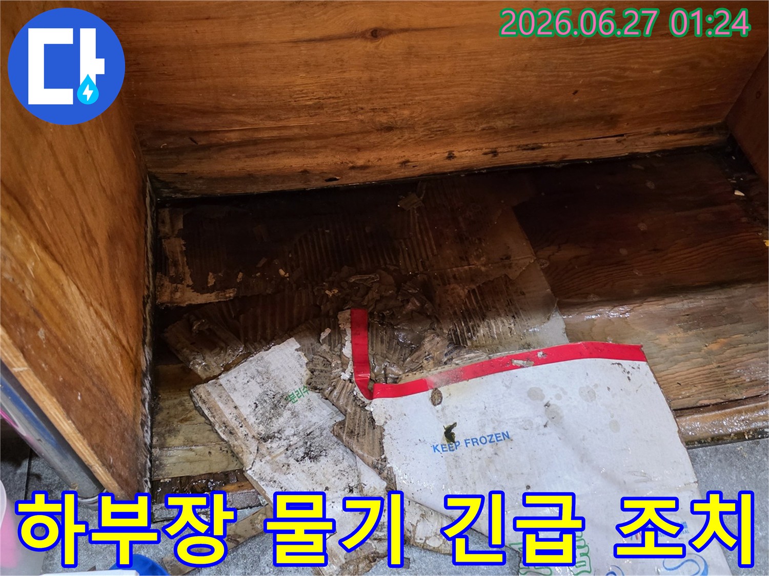 물에 젖어-심하게 손상된-종이 상자와-습기로 얼룩진-나무 바닥이-보이는-싱크대 하부장의-누수 현장입니다.