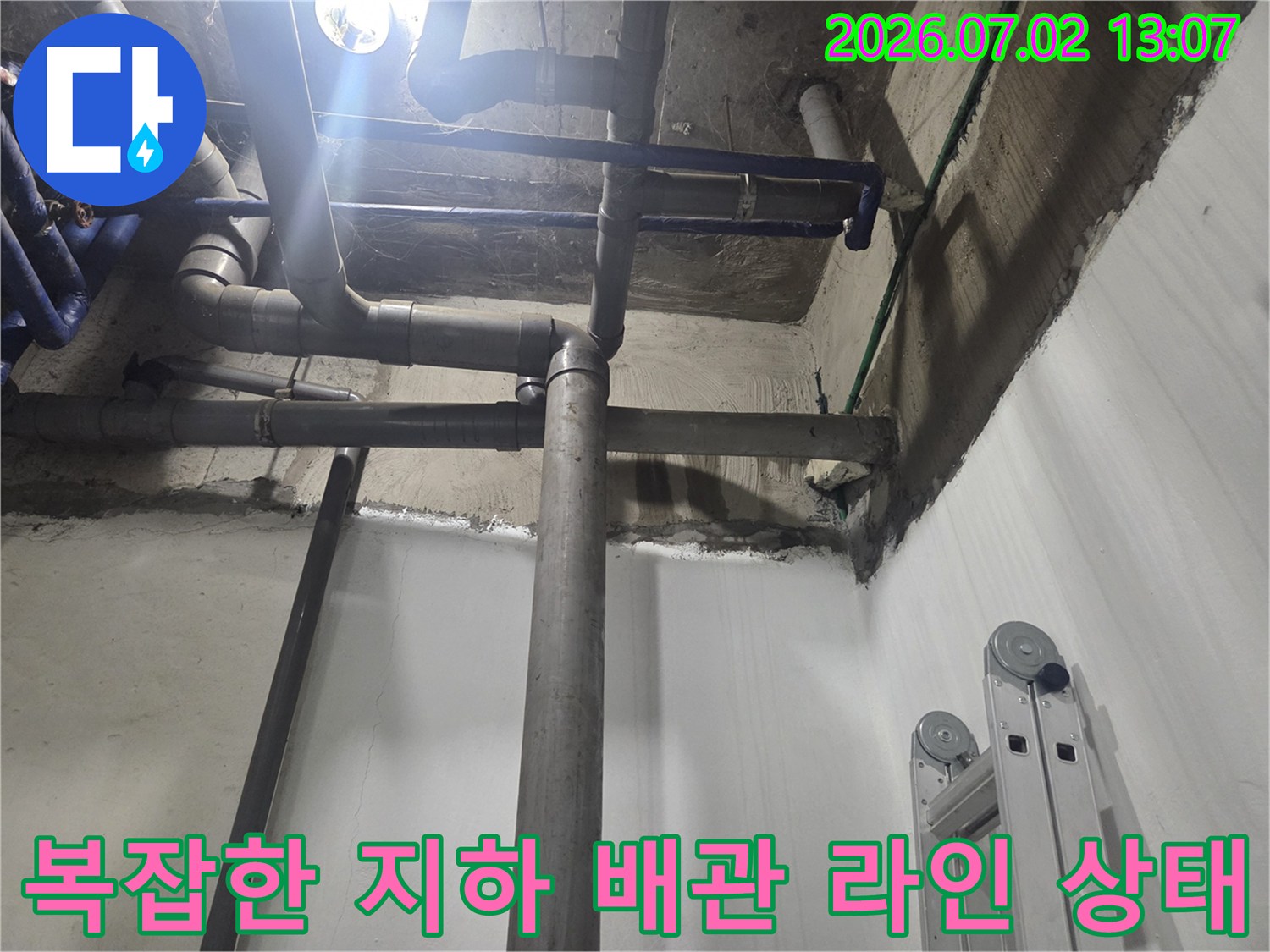 천장에서 수직으로 내려오는 회색 하수 배관-벽면의 젖은 흔적과 콘크리트 마감