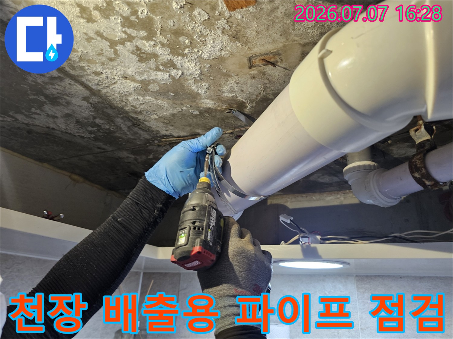 어두운 천장 내부에서 작업자가 손전등과 장비를 활용해 배수 파이프를 확인하는 모습