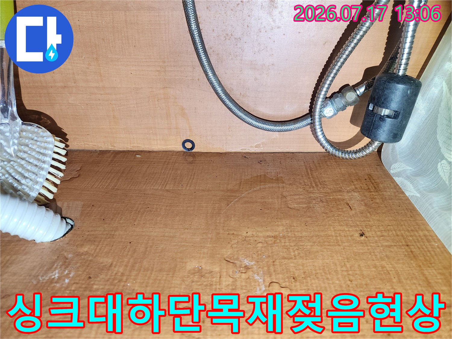 물에 젖어 어둡게 변한 주방 하부장 내부 나무 벽면-금속 배관과 흰색 호스의 연결 상태