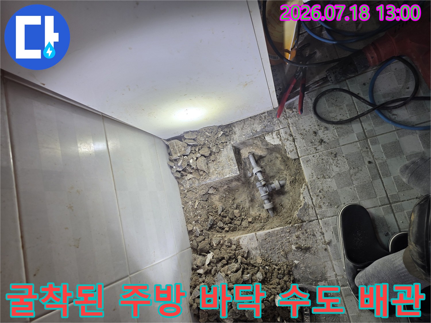 흰색 하부장 아래쪽의 타일을 제거하고 굴착하여 드러낸 금속제 수도 배관-누수 지점