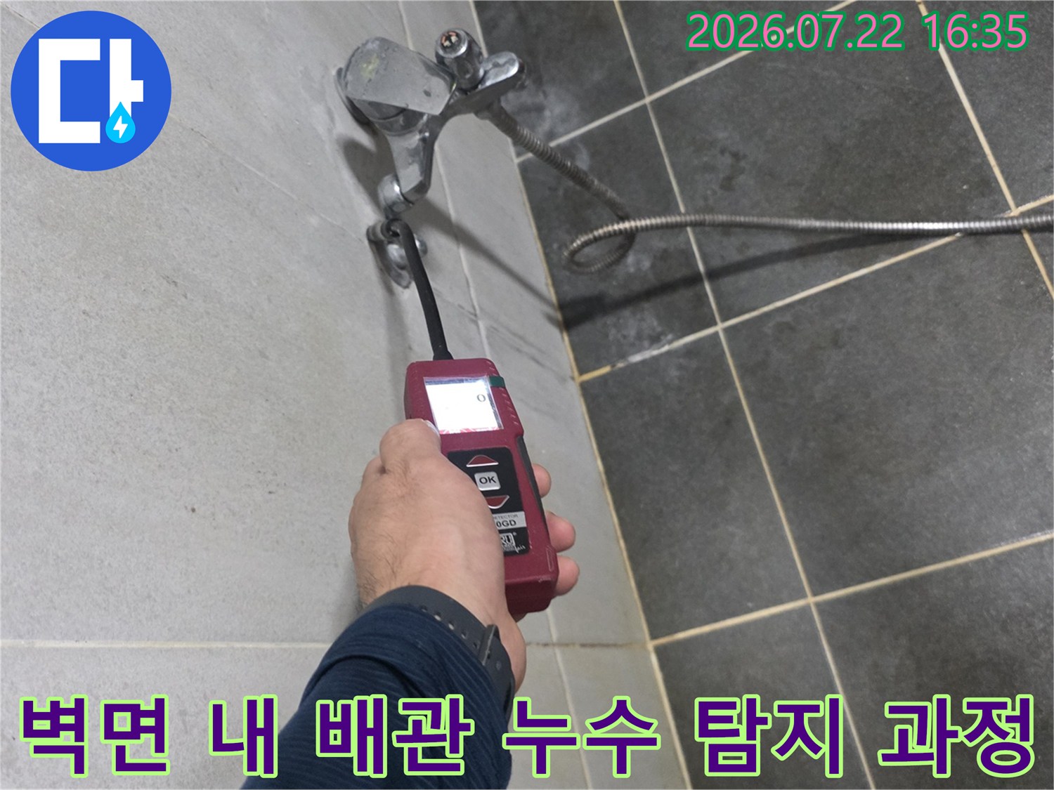 회색 타일 벽면과-금속 호스가 연결된 부위에서-디지털 측정기로-누수 여부를-체크하는 엔지니어의 손