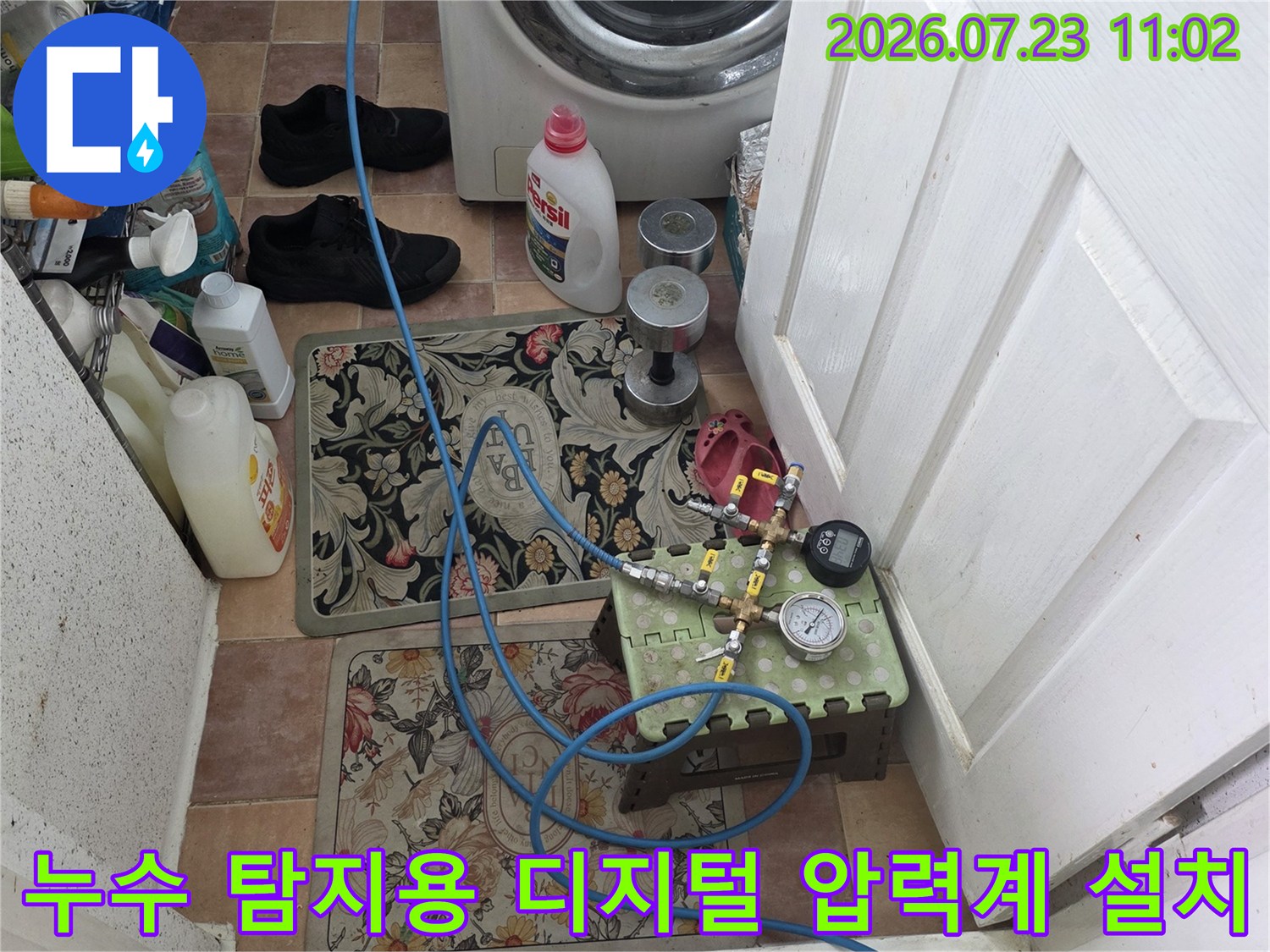 세탁기 옆 공간에 배치된 파란색 호스와 함께 작동 중인 디지털 압력 측정 장치