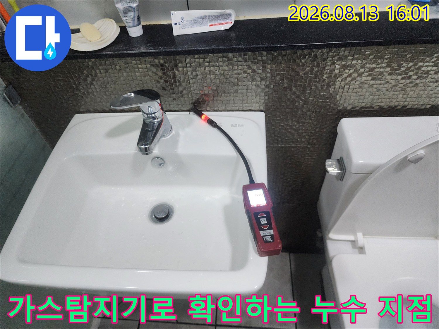 세면대 뒤쪽 벽면 배관 부위에서 가스 탐지기를 사용하여 누수 여부를 정밀하게 검사하고 있습니다.