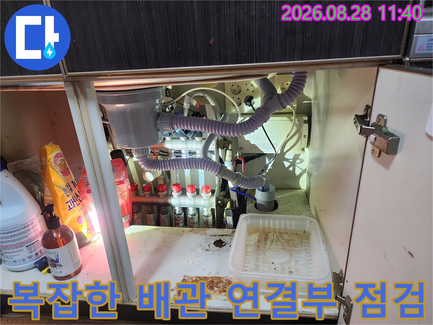 싱크대 하부장 내부의 정수기 필터와 에어컨 드레인 호스 및 배관들이 노출된 장면입니다.