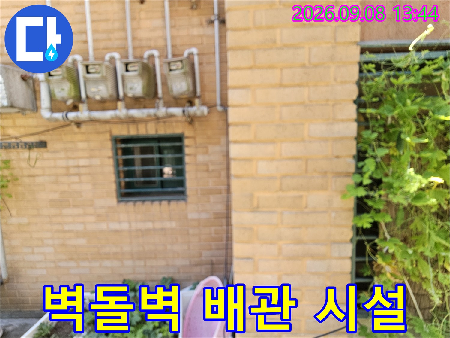 황색 벽돌 외벽에 설치된 수도 계량기와 상단 연결 배관입니다.