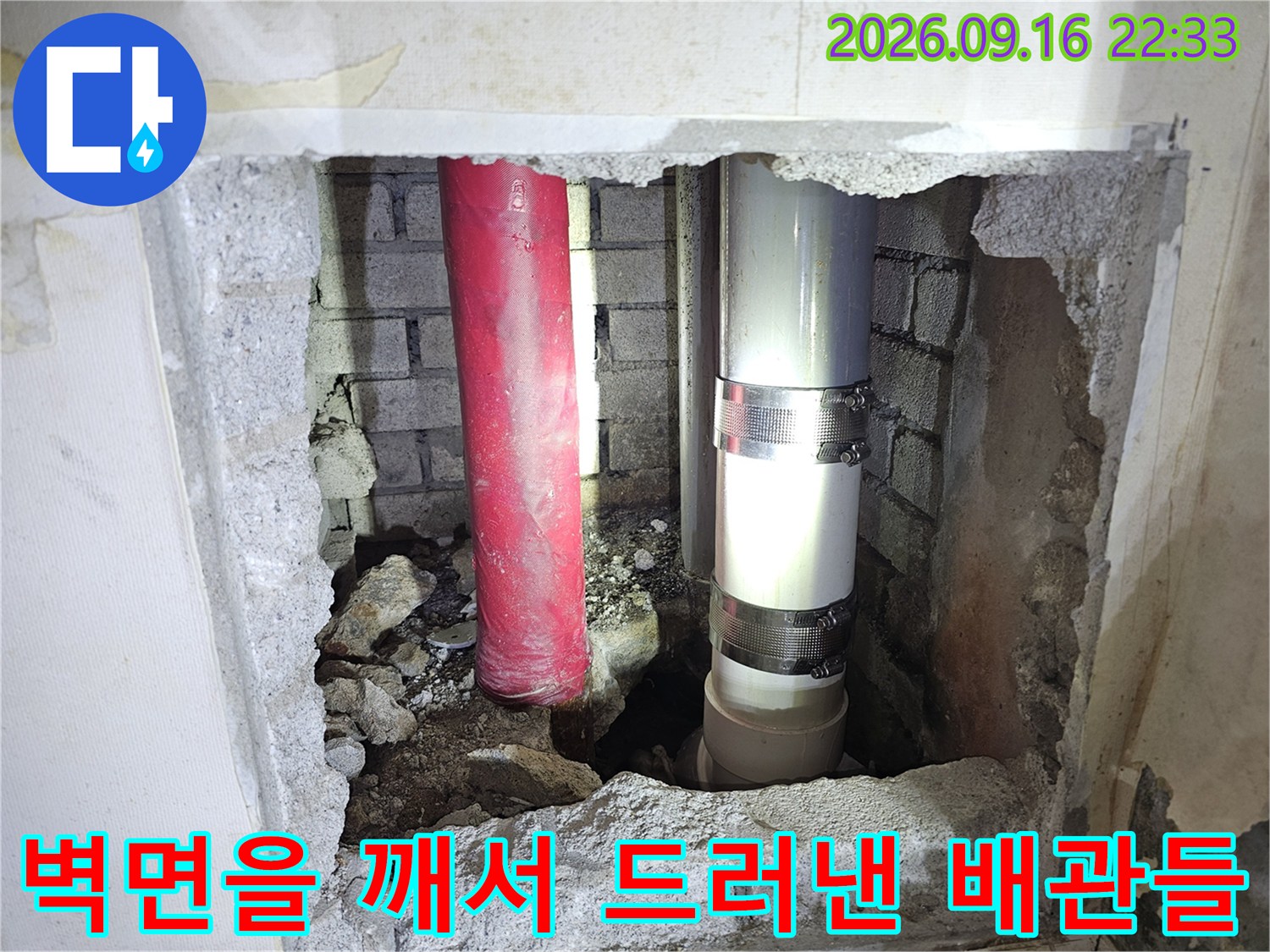 굴착된 벽 틈 사이로 보이는 빨간색 및 은색의 매립 배관입니다.
